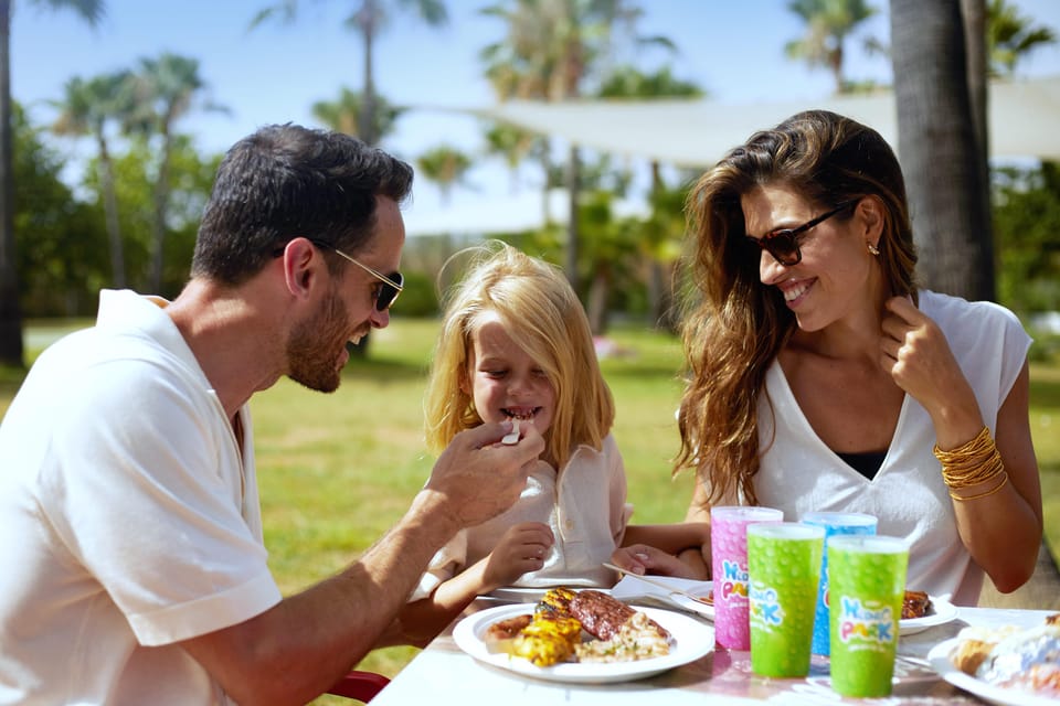 Happy family enjoying a delicious meal together at Hidropark Alcúdia, surrounded by palm trees and sunny skies.